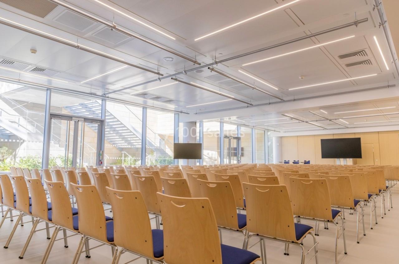 Salle de conférence lumineuse avec des rangées de chaises en bois, deux écrans et de grandes fenêtres donnant sur l…