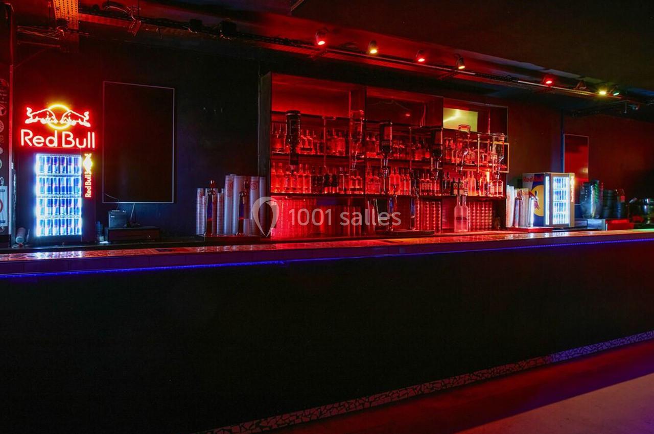 Bar sombre avec éclairage rouge, étagères remplies de bouteilles, et réfrigérateurs lumineux affichant le logo Red Bull.