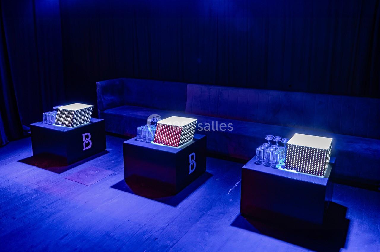 Tables lumineuses avec des cubes éclairés, entourées de verres, dans une salle sombre avec un canapé en arrière-plan.