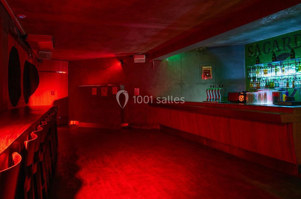 Salle de bar avec éclairage rouge tamisé, comptoir en bois à droite et rangée de tabourets à gauche.
