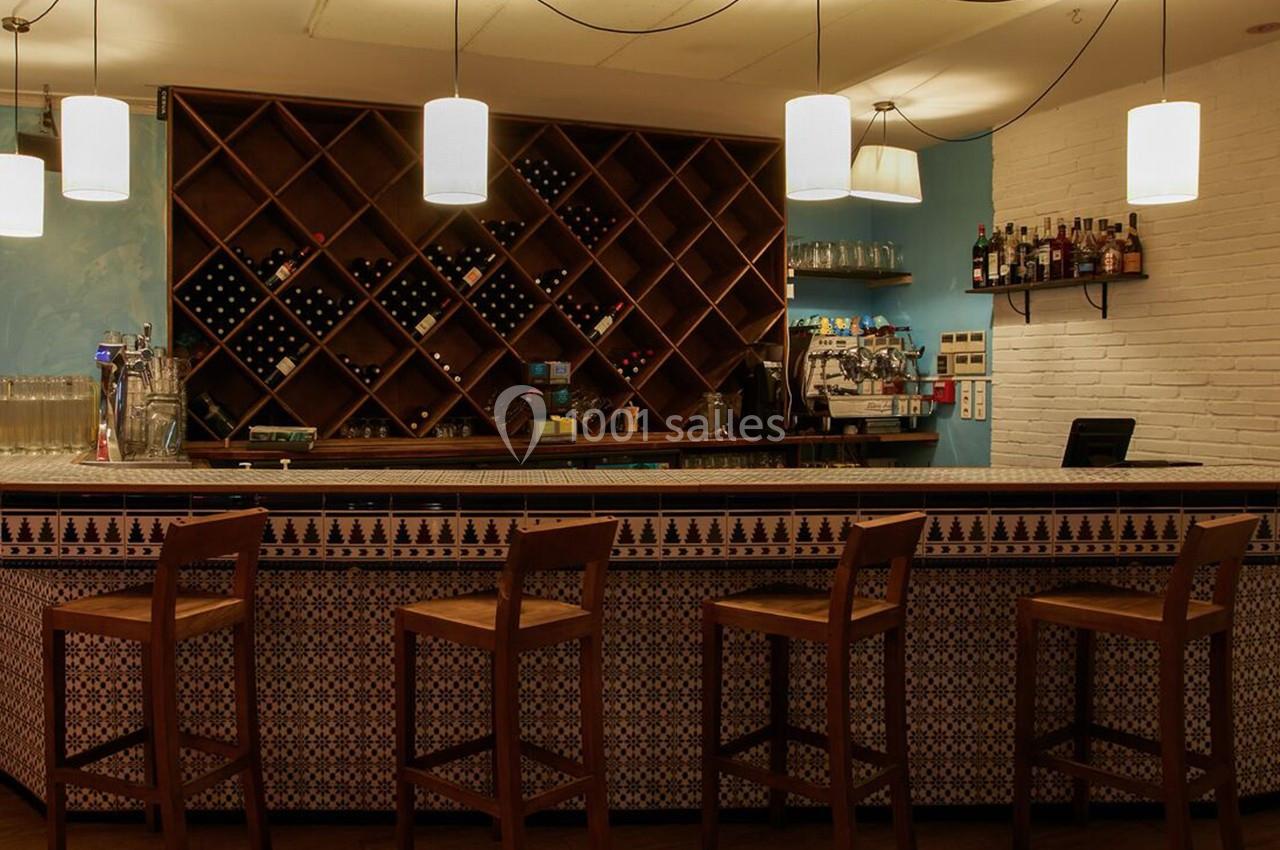 Bar avec comptoir carrelé, tabourets en bois, étagères de bouteilles et lumières suspendues dans une ambiance chaleureuse.