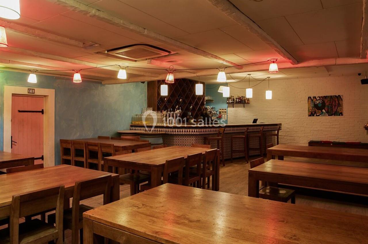 Salle de restaurant vide avec tables en bois, bar éclairé en arrière-plan et murs décorés de couleurs claires.
