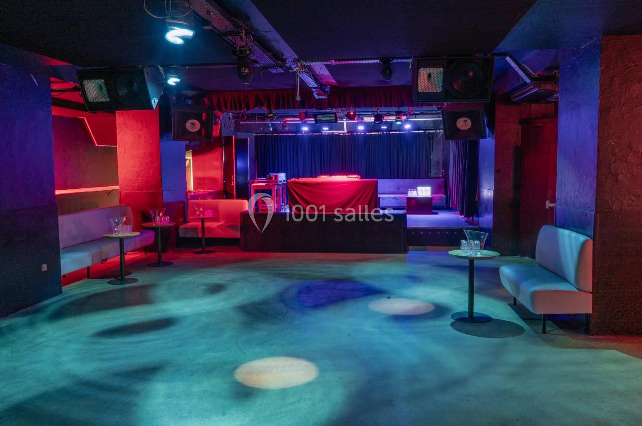 Salle de concert vide avec scène éclairée par des projecteurs colorés, entourée de banquettes et tables basses.