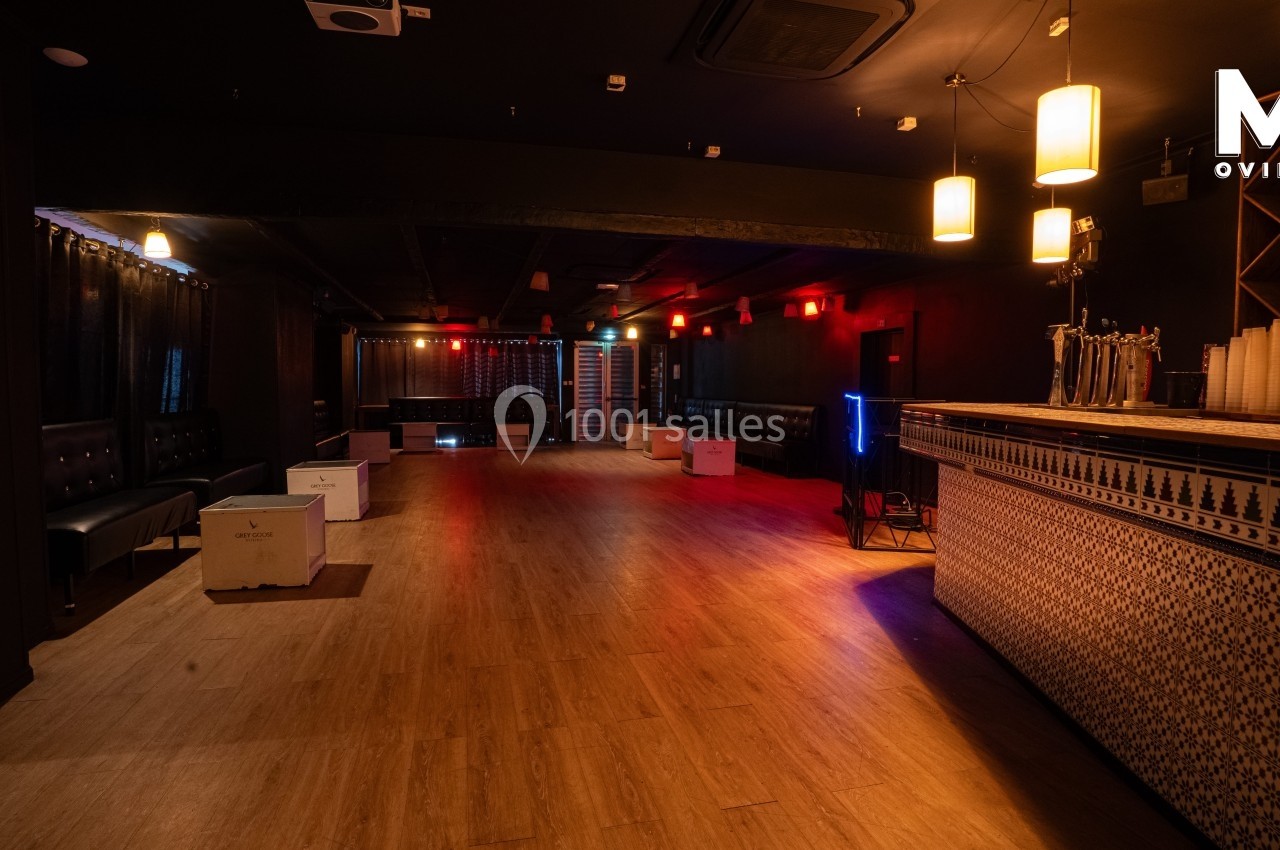 Salle sombre avec parquet, bar à droite, banquettes noires sur les côtés et éclairage tamisé.