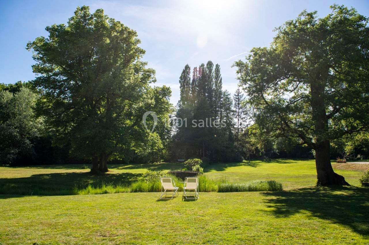 Deux chaises longues blanches sur une pelouse verdoyante entourée d'arbres sous un ciel ensoleillé.
