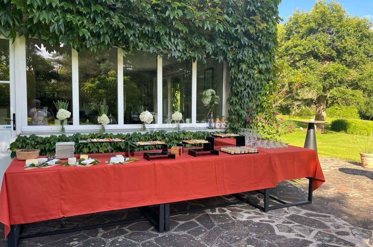 Table dressée avec une nappe rouge, contenant des plats, verres et décorations florales, en extérieur près d'un bâtiment…