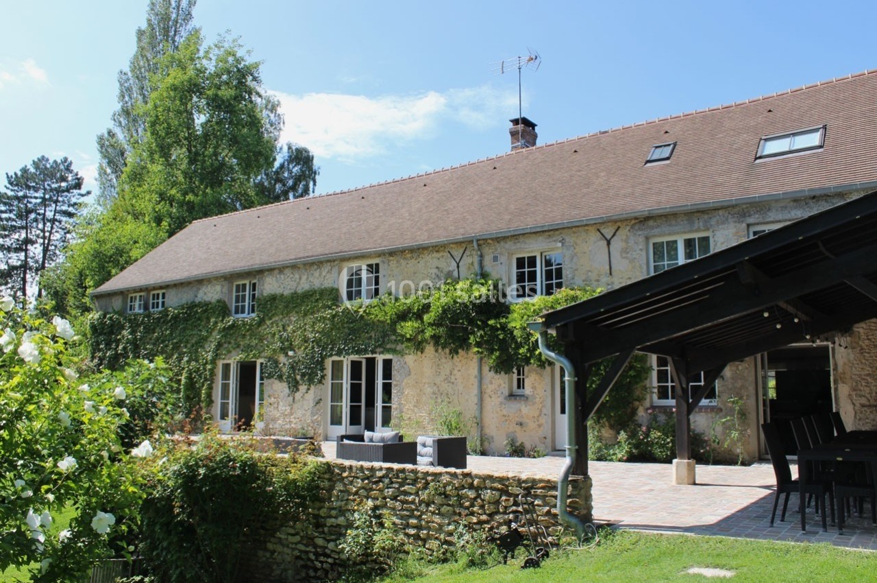 Façade d'une maison en pierre avec toit en tuiles, terrasse pavée, végétation grimpante et jardin arboré.