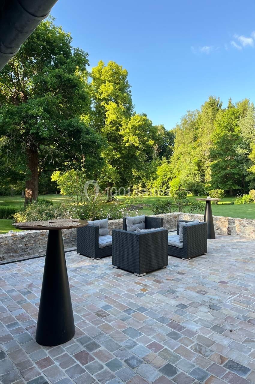 Mobilier de jardin en rotin noir avec coussins gris, disposé sur une terrasse en pierre, entourée de verdure.