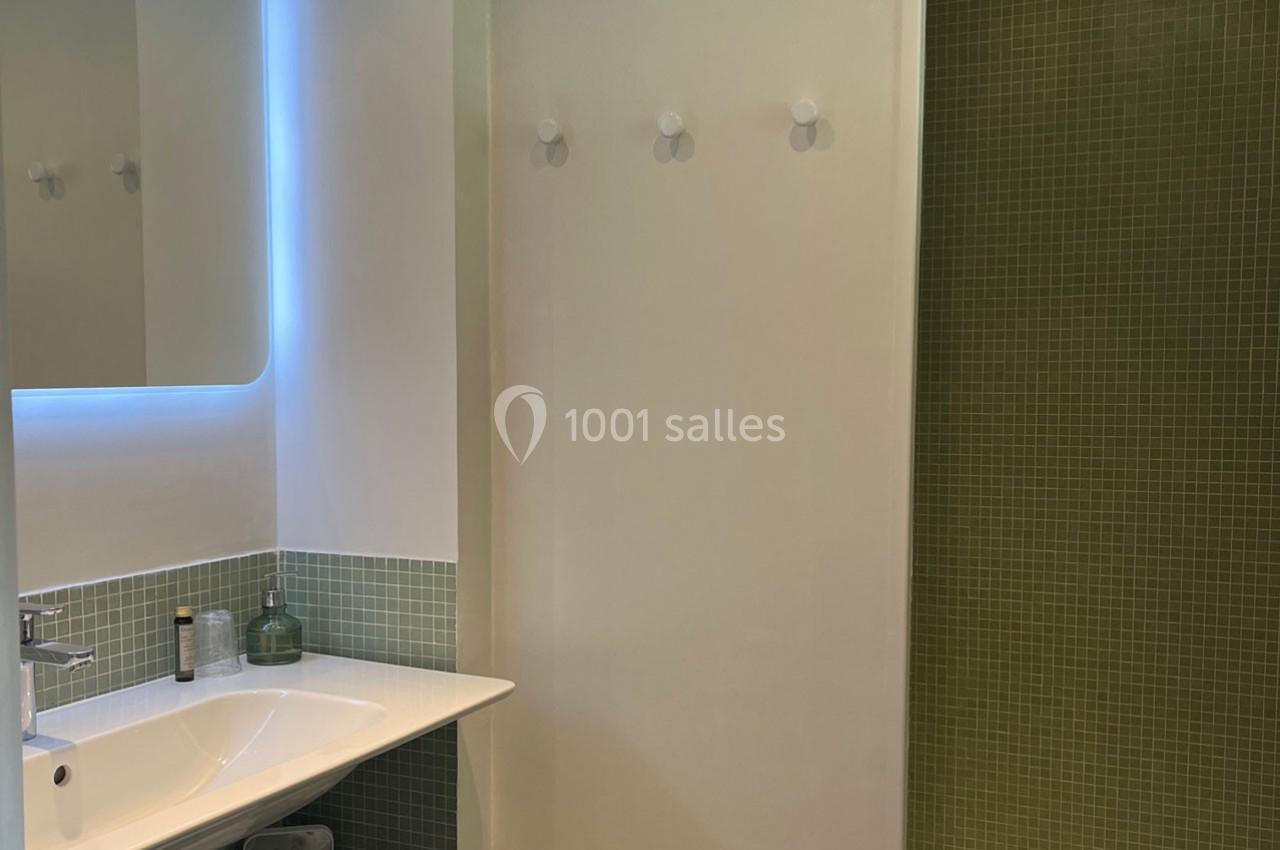 Salle de bain moderne avec lavabo, miroir éclairé, mur en carrelage vert et crochets muraux blancs.