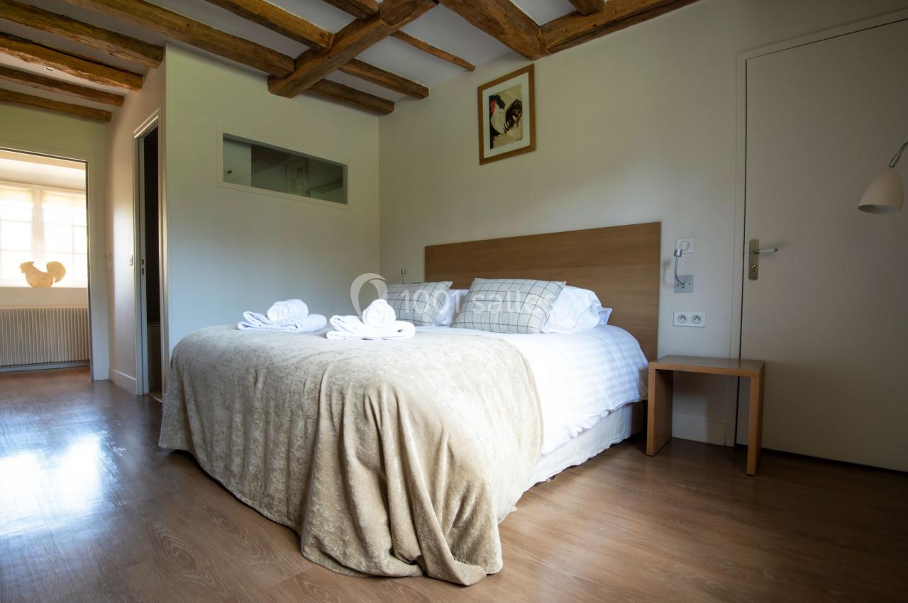 Chambre lumineuse avec lit double, draps blancs, poutres apparentes, sol en bois et décoration murale simple.