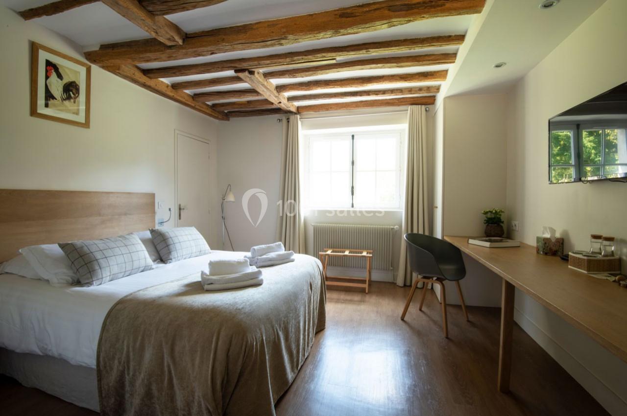 Chambre lumineuse avec lit double, poutres apparentes, bureau en bois, fenêtre donnant sur un extérieur verdoyant.