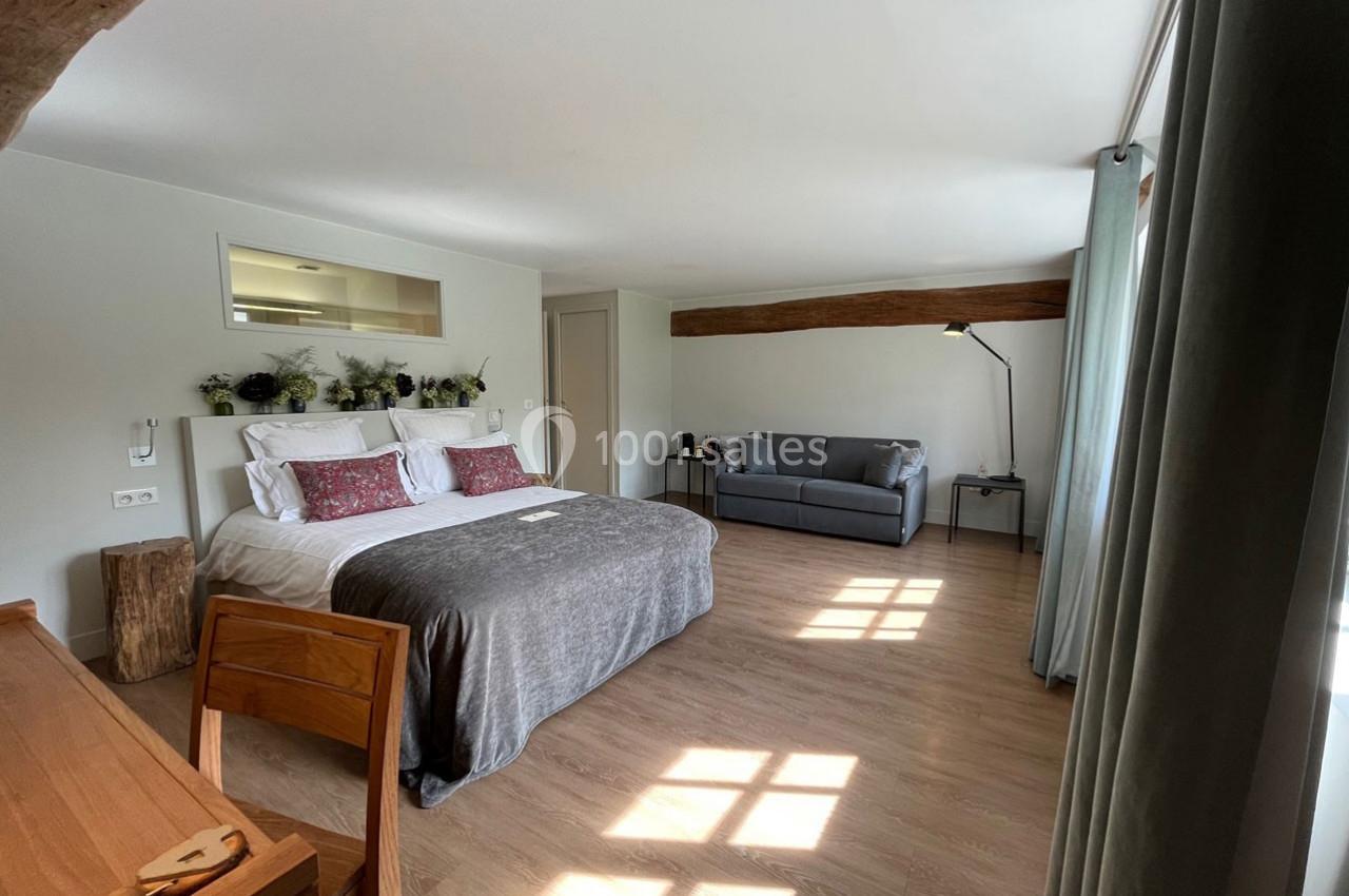 Chambre lumineuse avec lit double, canapé, bureau en bois, plantes décoratives et grande fenêtre laissant entrer la lumière.