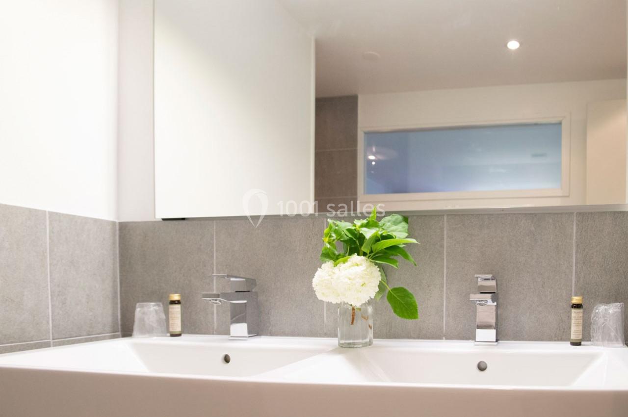 Double vasque avec robinetterie moderne, carrelage gris, miroir et vase contenant des fleurs blanches et vertes.