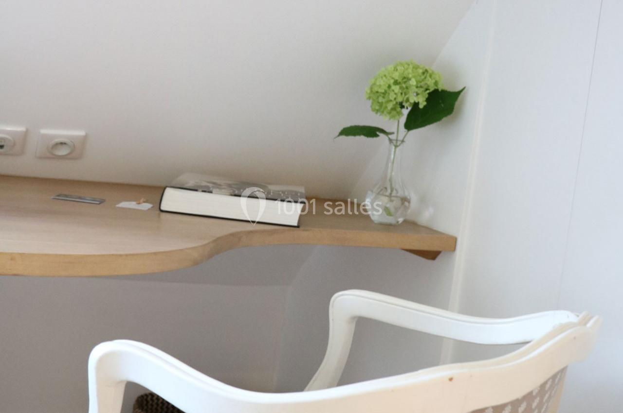 Table en bois clair avec un livre, une carte et un vase contenant une fleur verte, près d'un fauteuil blanc.