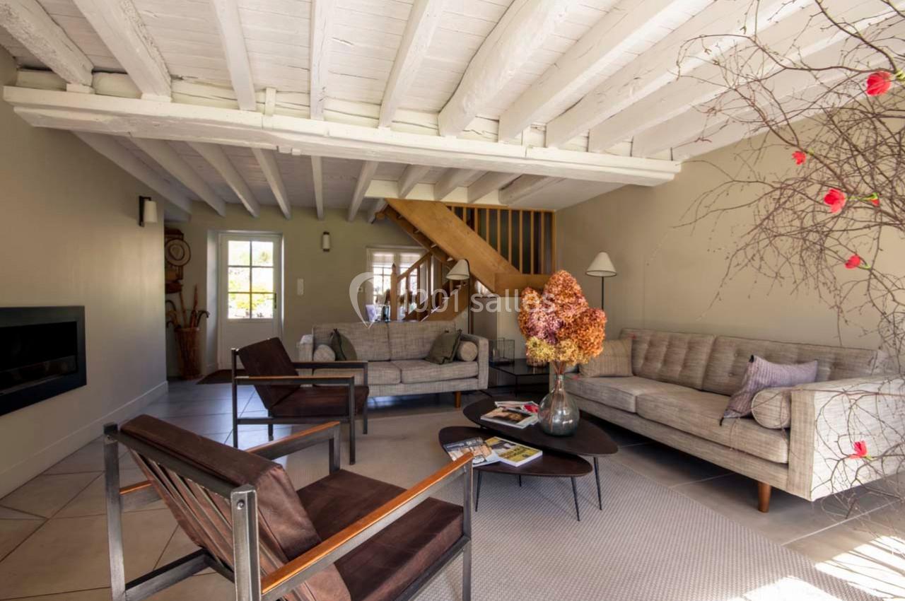 Salon lumineux avec poutres apparentes, canapé beige, fauteuils en bois, table basse et décoration florale.