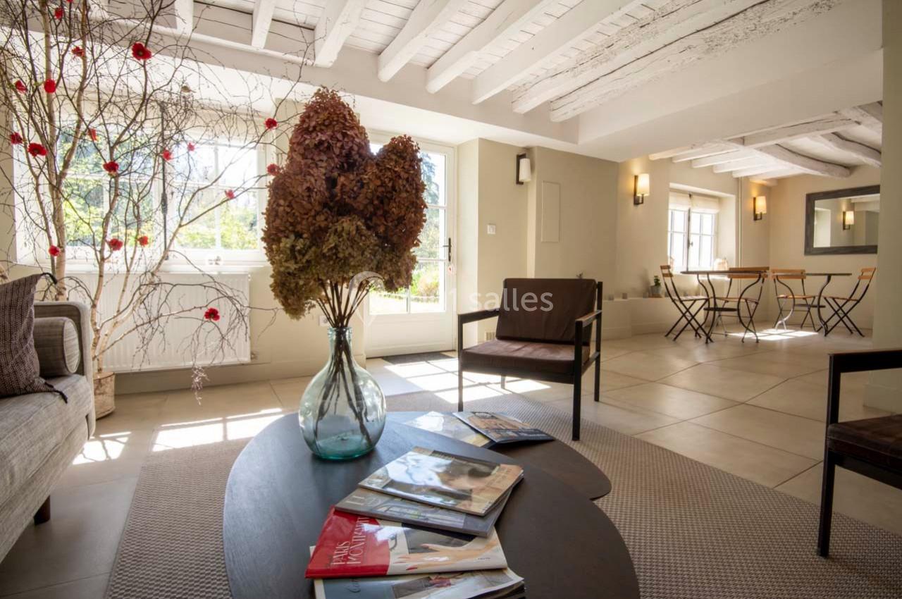 Salon lumineux avec poutres apparentes, table basse ornée de magazines et vase, vue sur une salle à manger.