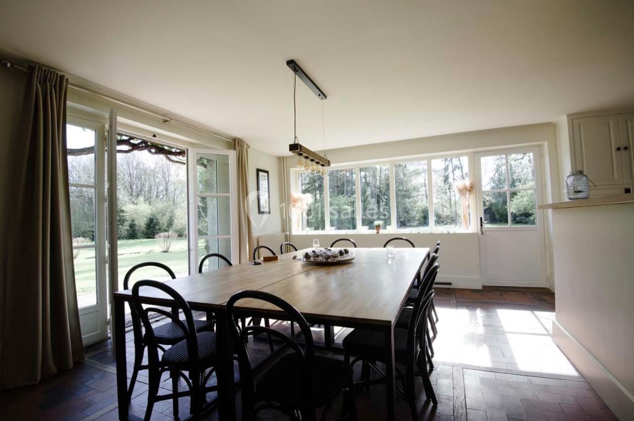Salle à manger lumineuse avec une grande table en bois entourée de chaises, donnant sur un jardin par des baies vitrées.