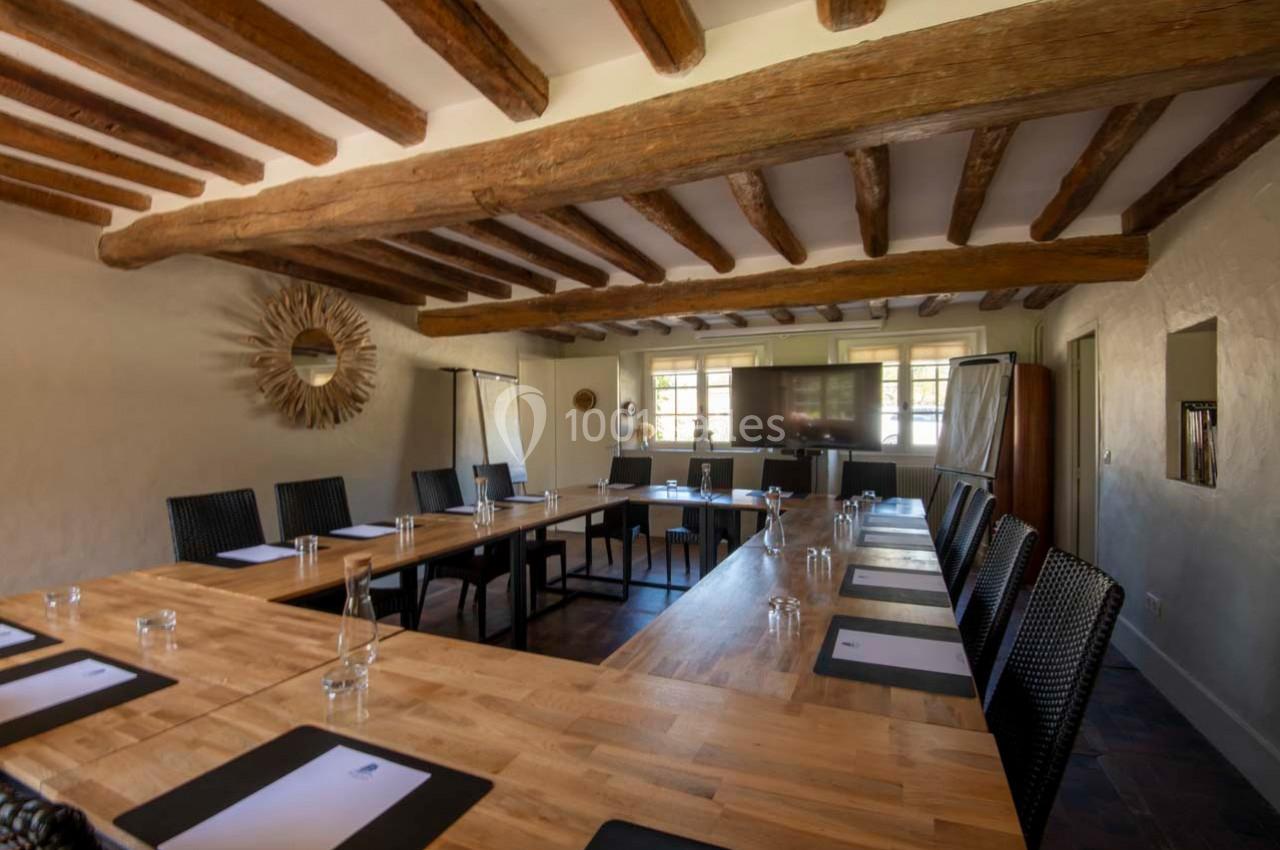 Salle de réunion lumineuse avec une grande table en bois, chaises noires, écran et poutres apparentes au plafond.