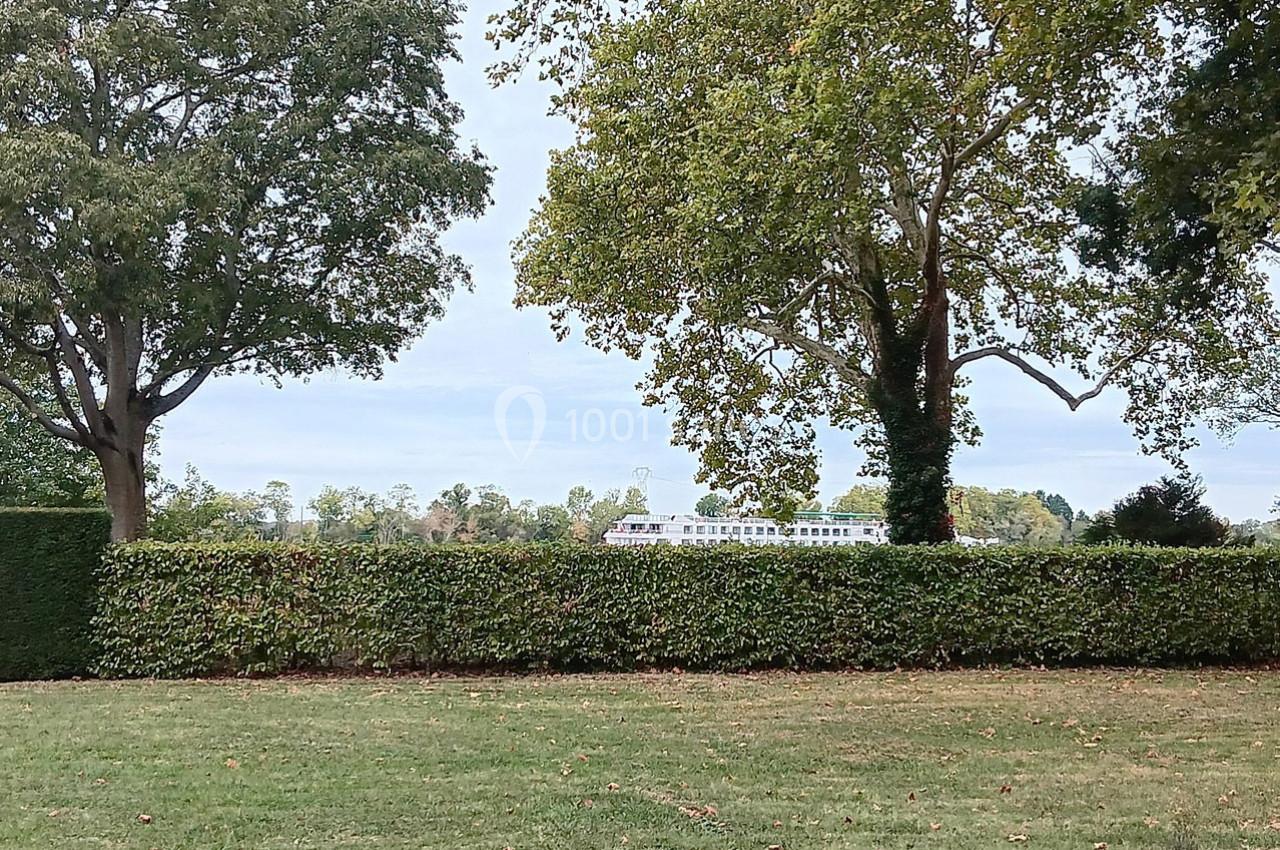 Vue d'une pelouse bordée de haies et d'arbres, avec un bâtiment blanc visible en arrière-plan.