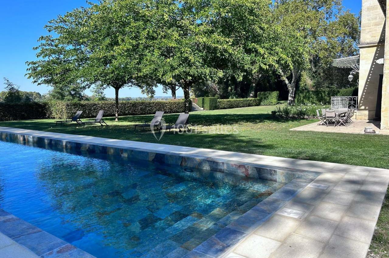 Piscine extérieure bordée de dalles claires, entourée de chaises longues, pelouse et arbres sous un ciel bleu.