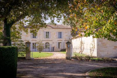 Façade d'une grande maison en pierre claire avec volets bleus, entourée d'un jardin et d'une allée gravillonnée.