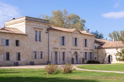 Façade d'une grande maison en pierre claire avec volets bleus, entourée d'un jardin et d'une allée gravillonnée.