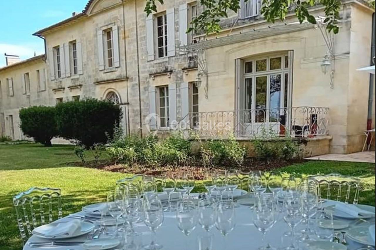 Table dressée avec verres et assiettes dans un jardin, devant une grande bâtisse en pierre avec volets blancs.