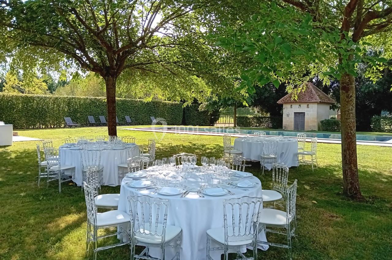 Tables rondes dressées avec nappes blanches sous des arbres dans un jardin, près d'une piscine et d'un petit bâtiment.