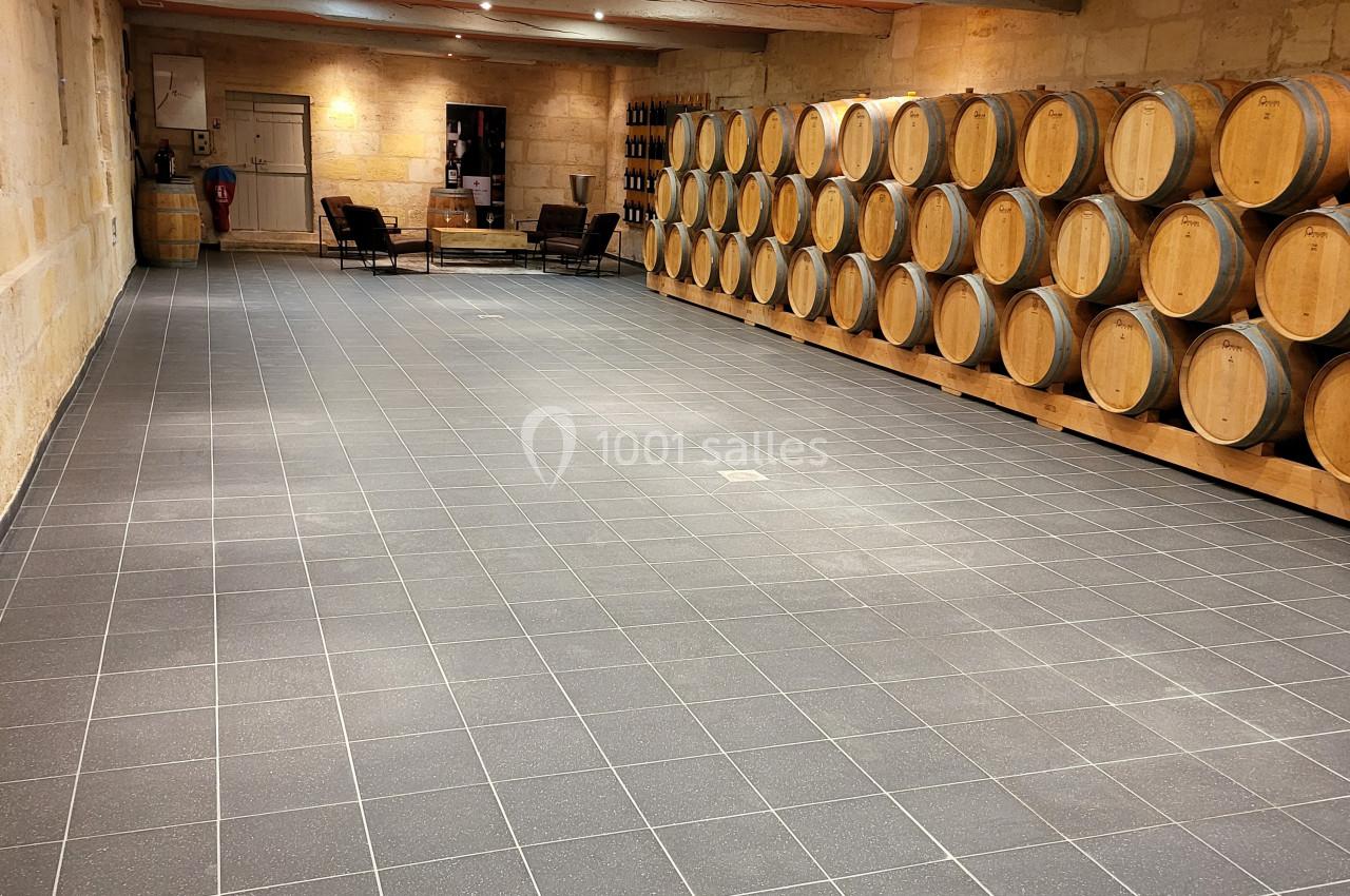 Salle de stockage avec des rangées de tonneaux en bois alignés contre un mur en pierre, éclairée par des spots au plafond.