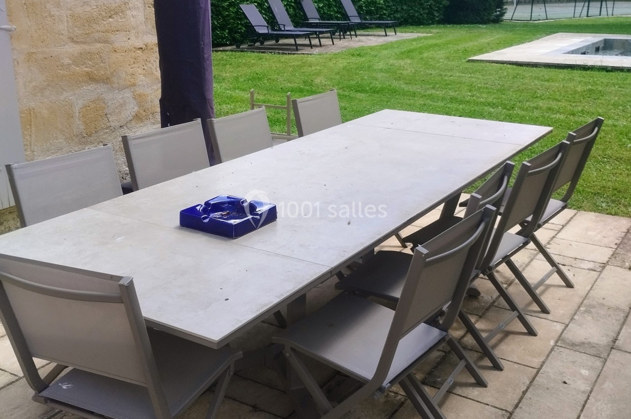 Table rectangulaire grise entourée de chaises pliantes sur une terrasse en pierre, avec jardin, piscine et transats en…