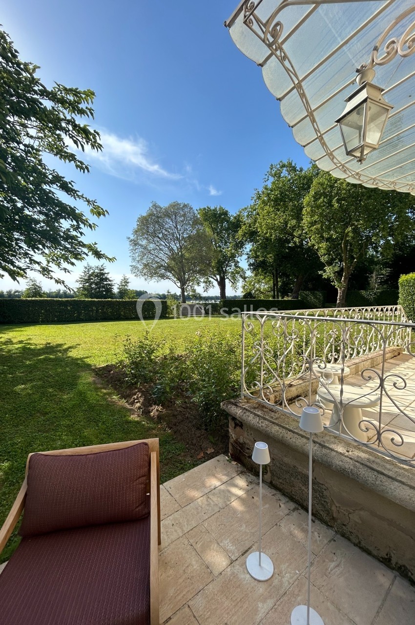 Terrasse en pierre avec fauteuil, lampes et balustrade, donnant sur un jardin arboré sous un ciel bleu dégagé.