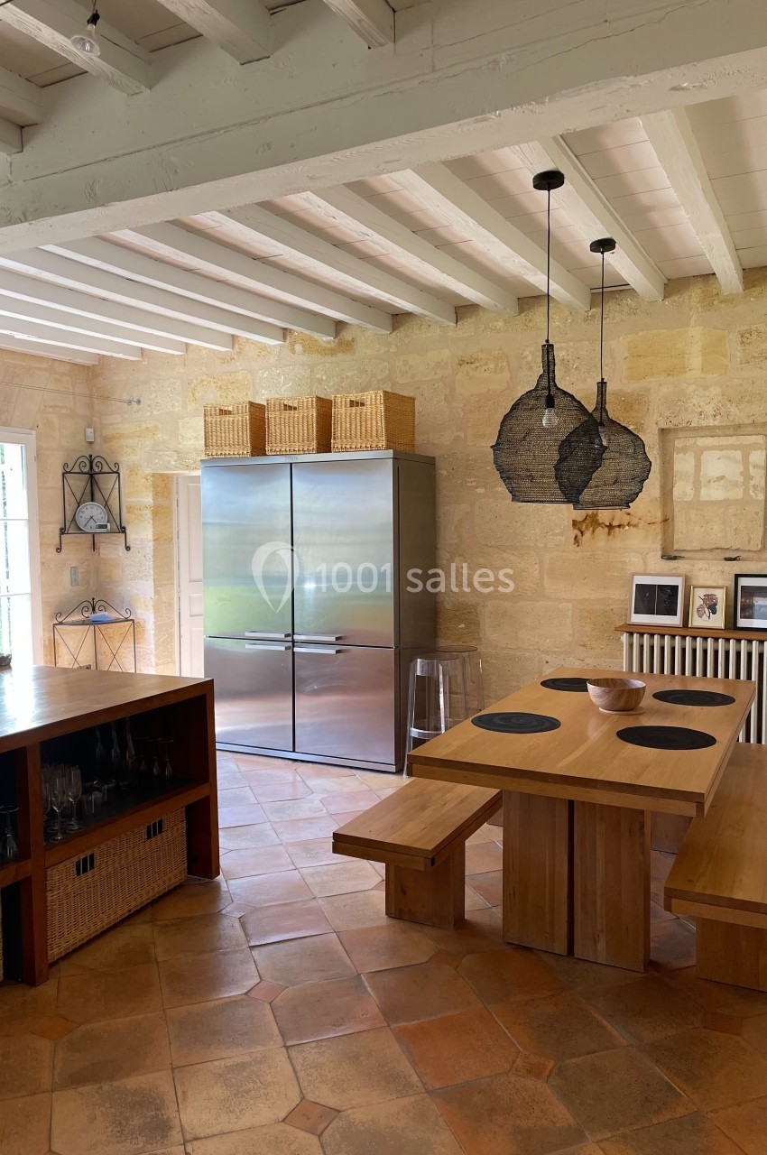 Cuisine lumineuse avec murs en pierre, table en bois, réfrigérateur en inox et suspensions noires modernes.