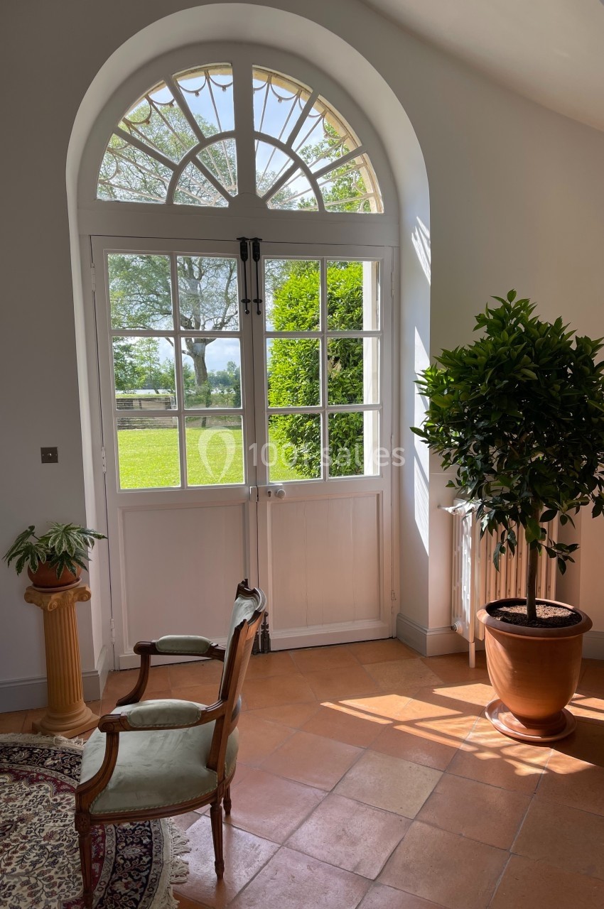 Porte-fenêtre cintrée donnant sur un jardin, éclairant un intérieur avec fauteuils, tapis et plante en pot.