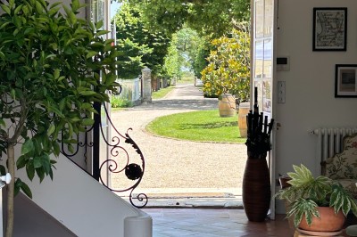 Façade d'une grande maison en pierre claire avec volets bleus, entourée d'un jardin et d'une allée gravillonnée.