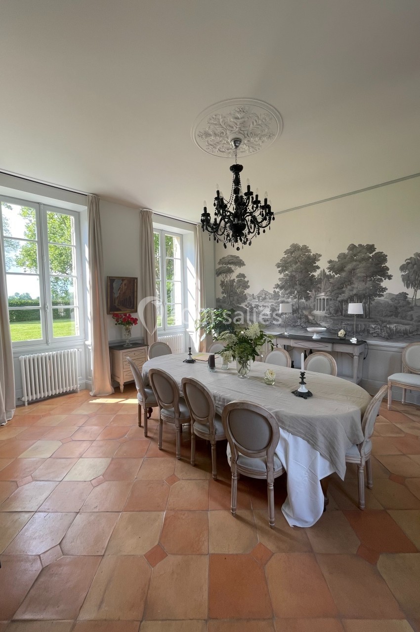 Salle à manger lumineuse avec une grande table, des chaises élégantes, un lustre noir et une fresque murale représentant un…