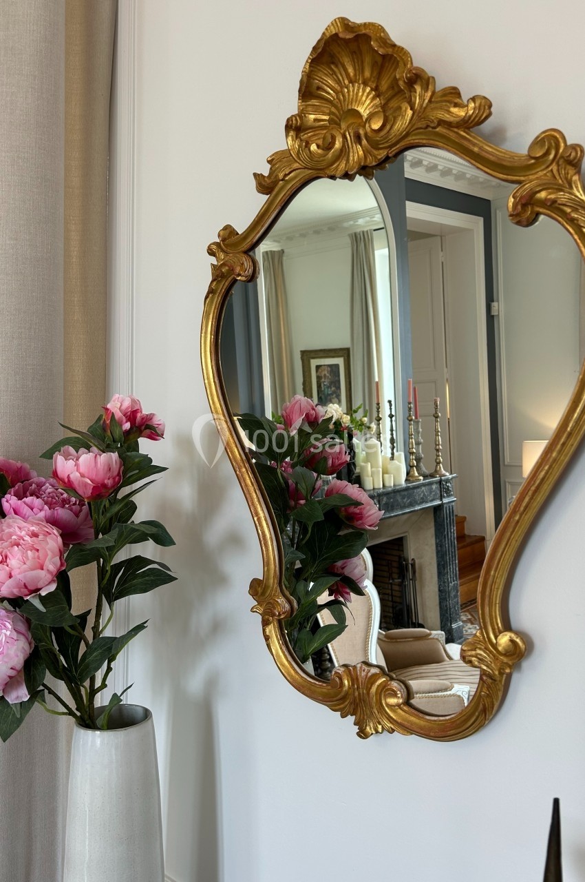 Miroir doré au cadre sculpté reflétant un intérieur élégant avec cheminée, bougies et rideaux beiges, vase de pivoines roses…