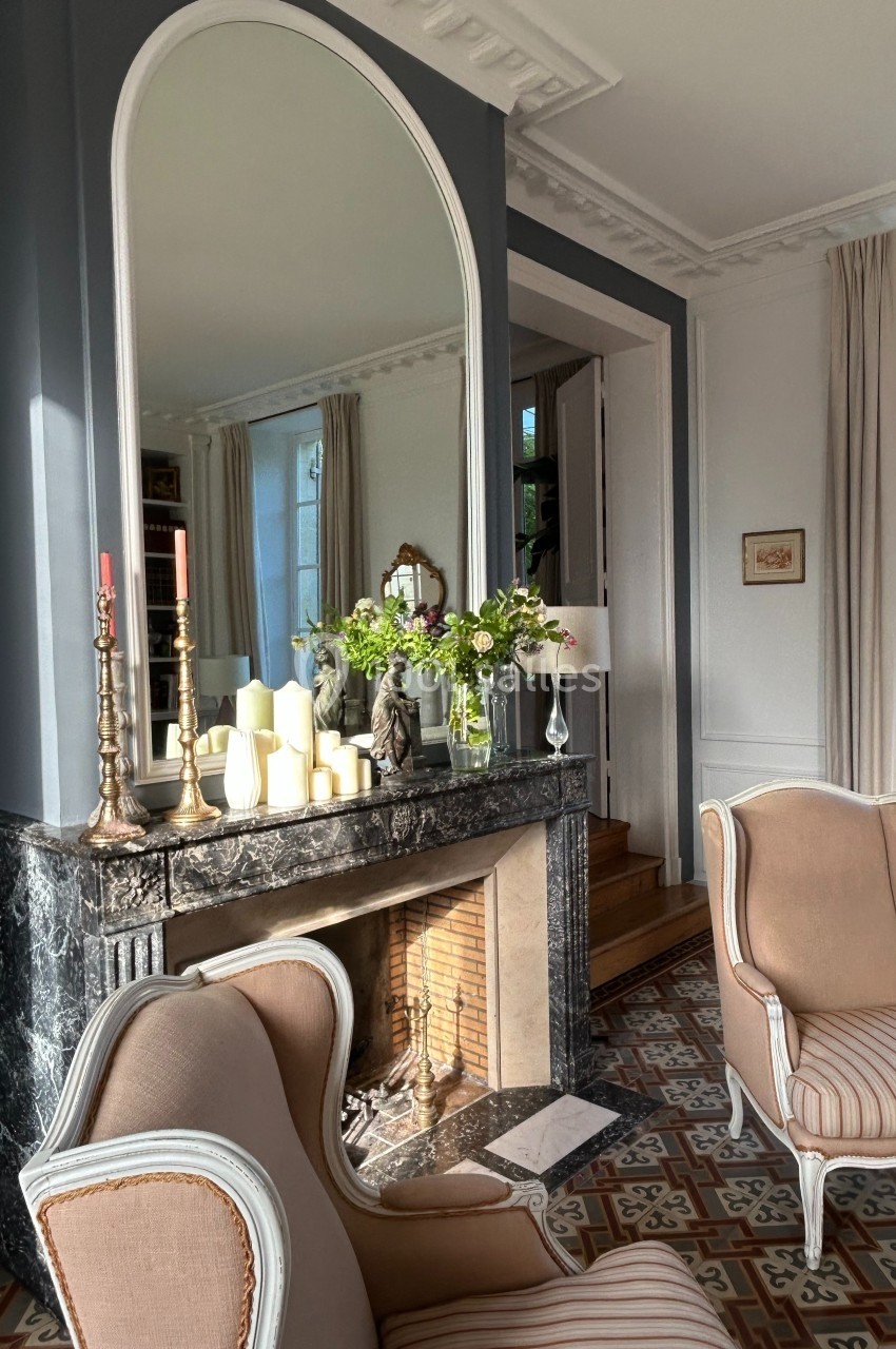 Cheminée en marbre noir décorée de bougies et de fleurs, entourée de fauteuils dans un salon lumineux.