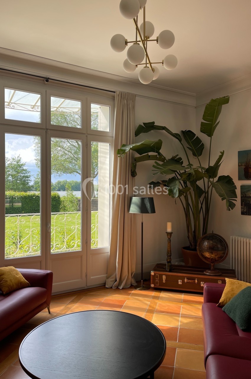 Salon lumineux avec grandes baies vitrées, mobilier moderne, plantes décoratives et vue sur un jardin verdoyant.