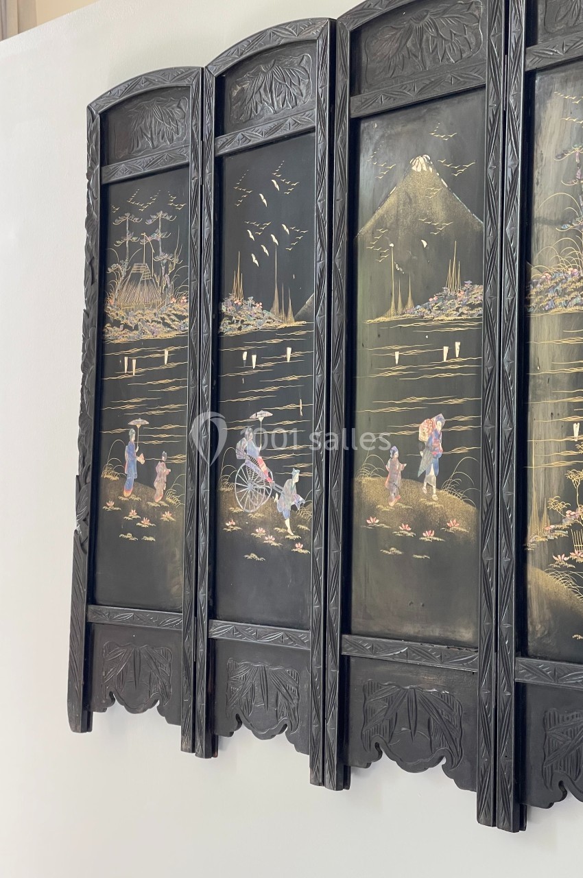 Paravent en bois noir orné de scènes gravées représentant des paysages et des personnages dans un décor asiatique.