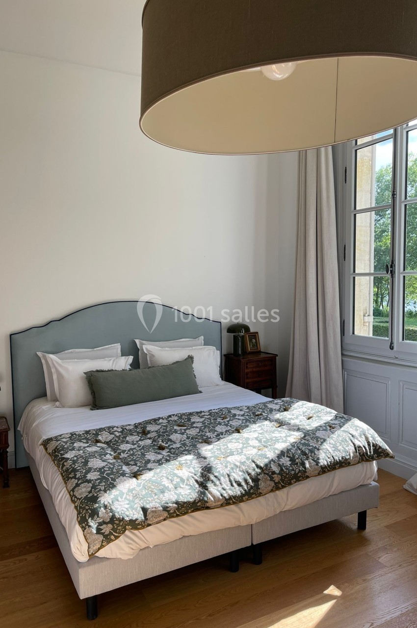 Chambre lumineuse avec un lit double, une grande fenêtre donnant sur un jardin et un luminaire suspendu.