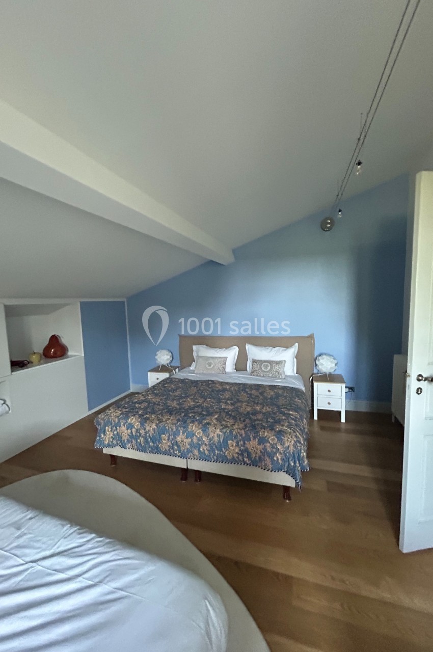 Chambre avec un lit double, mur bleu, plafond mansardé, parquet clair et étagères intégrées sur le côté.