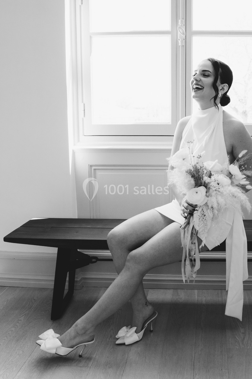 Une femme en robe blanche, assise sur un banc près d'une fenêtre, tenant un bouquet de fleurs, sourit joyeusement.