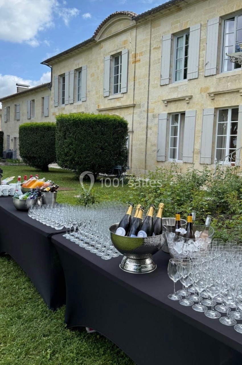 Tables dressées avec des verres et des bouteilles de champagne devant un bâtiment en pierre entouré de verdure.