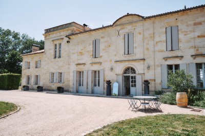 Façade d'une grande maison en pierre claire avec volets bleus, entourée d'un jardin et d'une allée gravillonnée.
