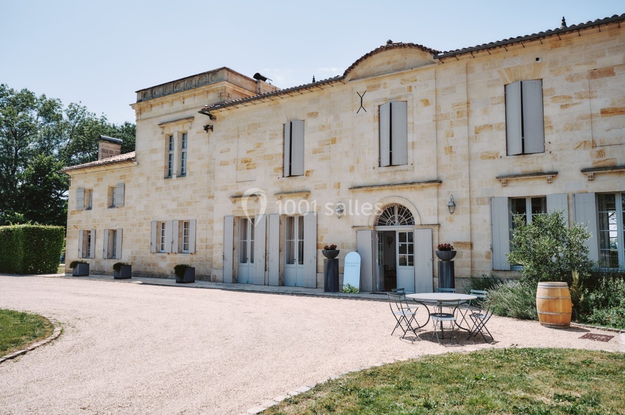 Façade d'une grande maison en pierre claire avec volets bleus, entourée d'un jardin et d'une allée gravillonnée.