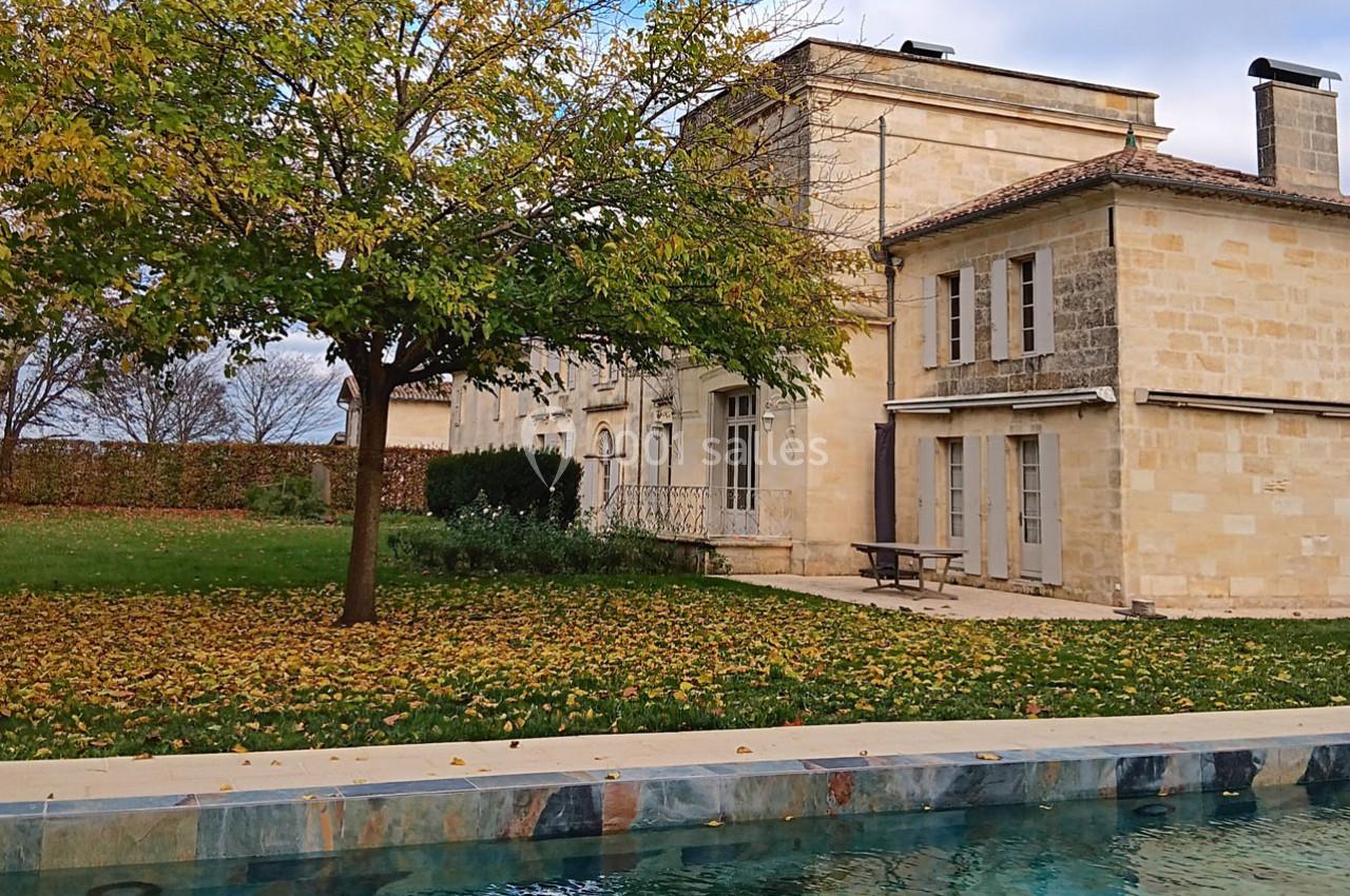 Maison en pierre entourée d'un jardin avec un arbre aux feuilles d'automne, vue depuis une piscine.