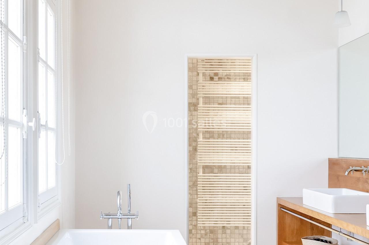 Salle de bain lumineuse avec baignoire blanche, meuble en bois, lavabo double et porte ouverte sur un sauna.