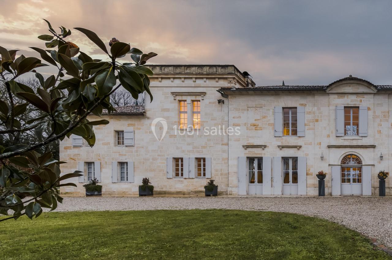Façade d'une maison en pierre claire avec fenêtres éclairées, entourée d'un jardin et d'un ciel nuageux au crépuscule.