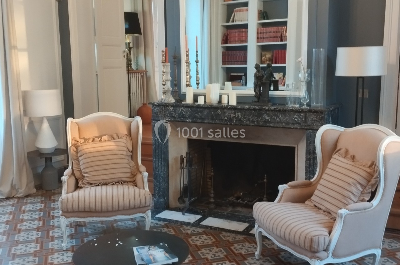 Deux fauteuils beiges devant une cheminée en marbre, entourés de bougies et d'étagères remplies de livres.