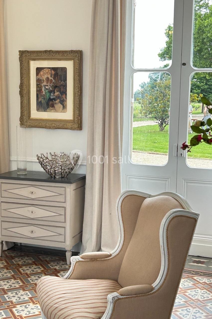 Fauteuil beige devant une commode grise, près d'une fenêtre ouverte donnant sur un jardin verdoyant.